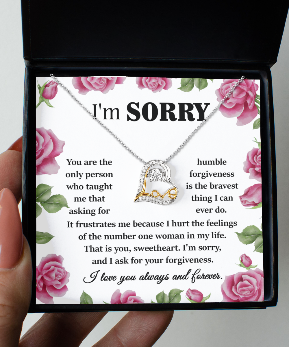 A person holds an open jewelry box containing the Sorry-Your Forgiveness - Love Dancing Necklace, a 14k gold heart-shaped pendant. The box features a heartfelt apology message surrounded by pink roses, expressing regret and asking for forgiveness.