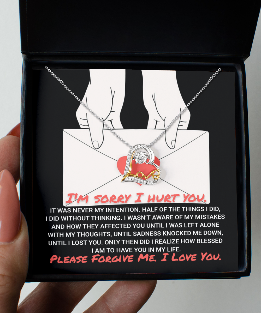 A hand holds an open box containing a Sorry-Without Thinking - Love Dancing Necklace, featuring a heart-shaped pendant crafted in sterling silver, resting on an envelope with an apology message printed below it.