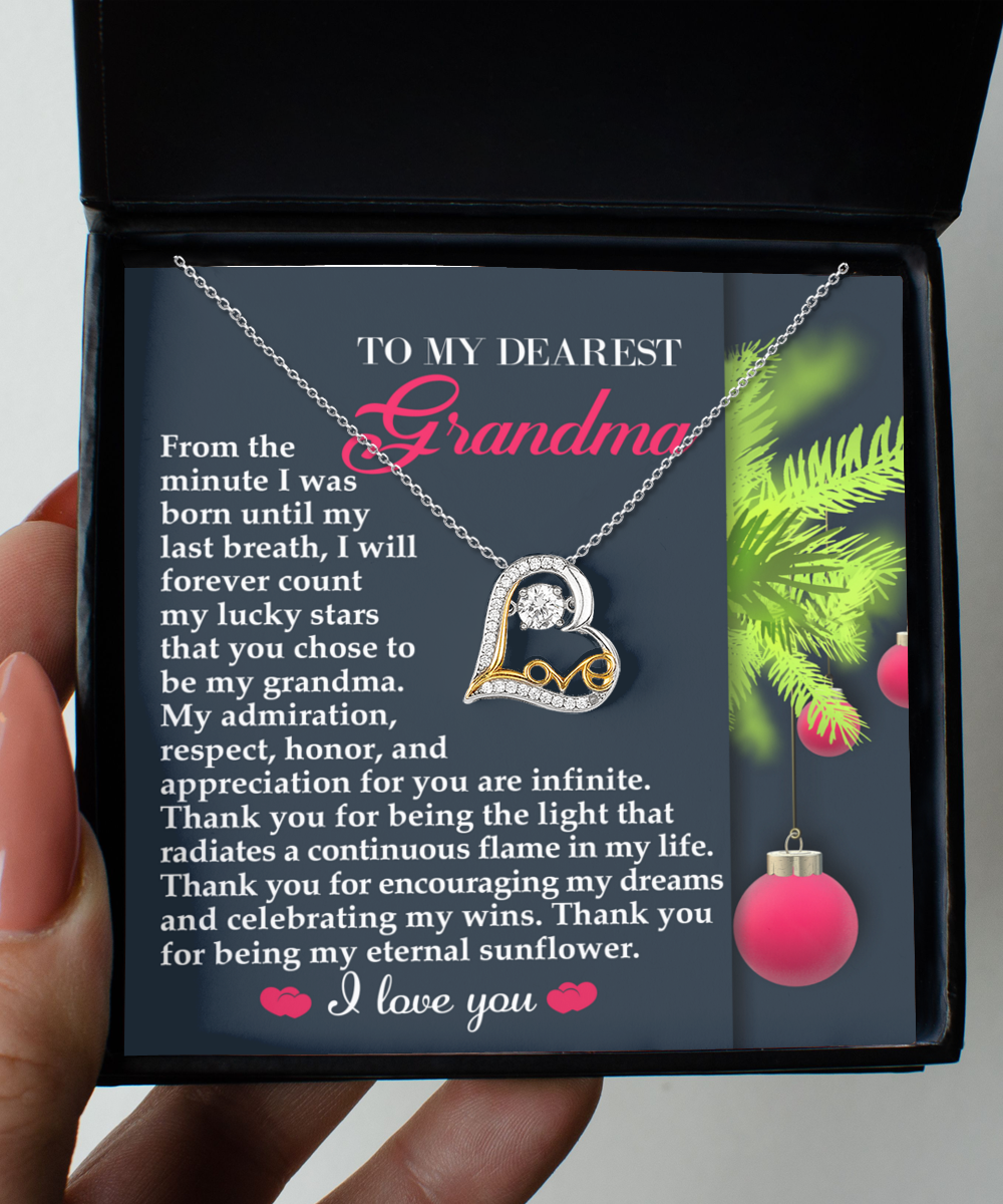 The Grandma-Eternal Sunflower - Love Dancing Necklace features a heart pendant on a gray card, expressing gratitude and admiration for Grandma. With a holiday theme, it illustrates maternal love with a green branch and red ornament.