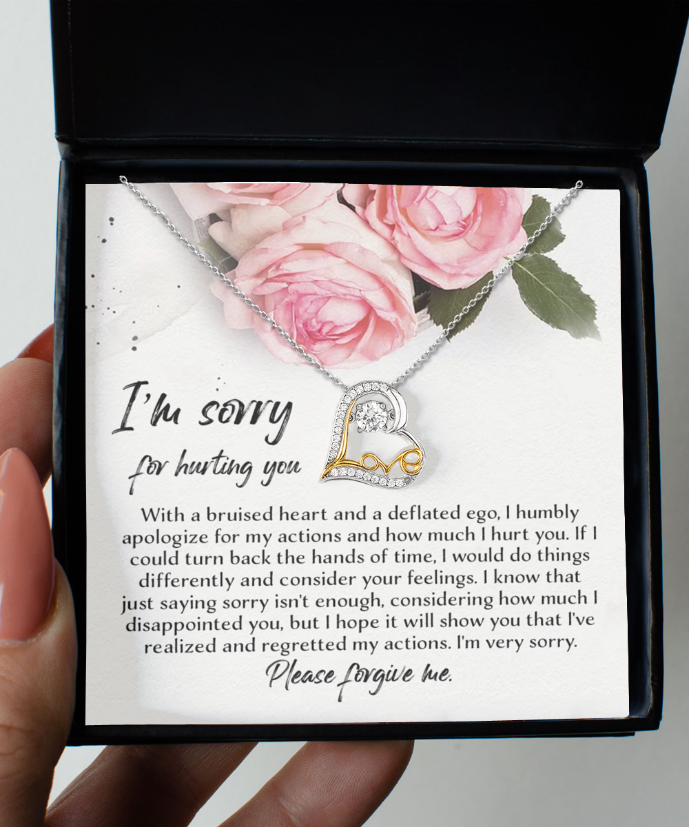 A hand holds an open jewelry box displaying the "Sorry-Regretted My Actions - Love Dancing Necklace," a heart-shaped pendant adorned with smaller hearts in gold and encrusted with Cubic Zirconia on a .925 Sterling Silver chain. The box includes a heartfelt apology message with pink roses printed at the top.