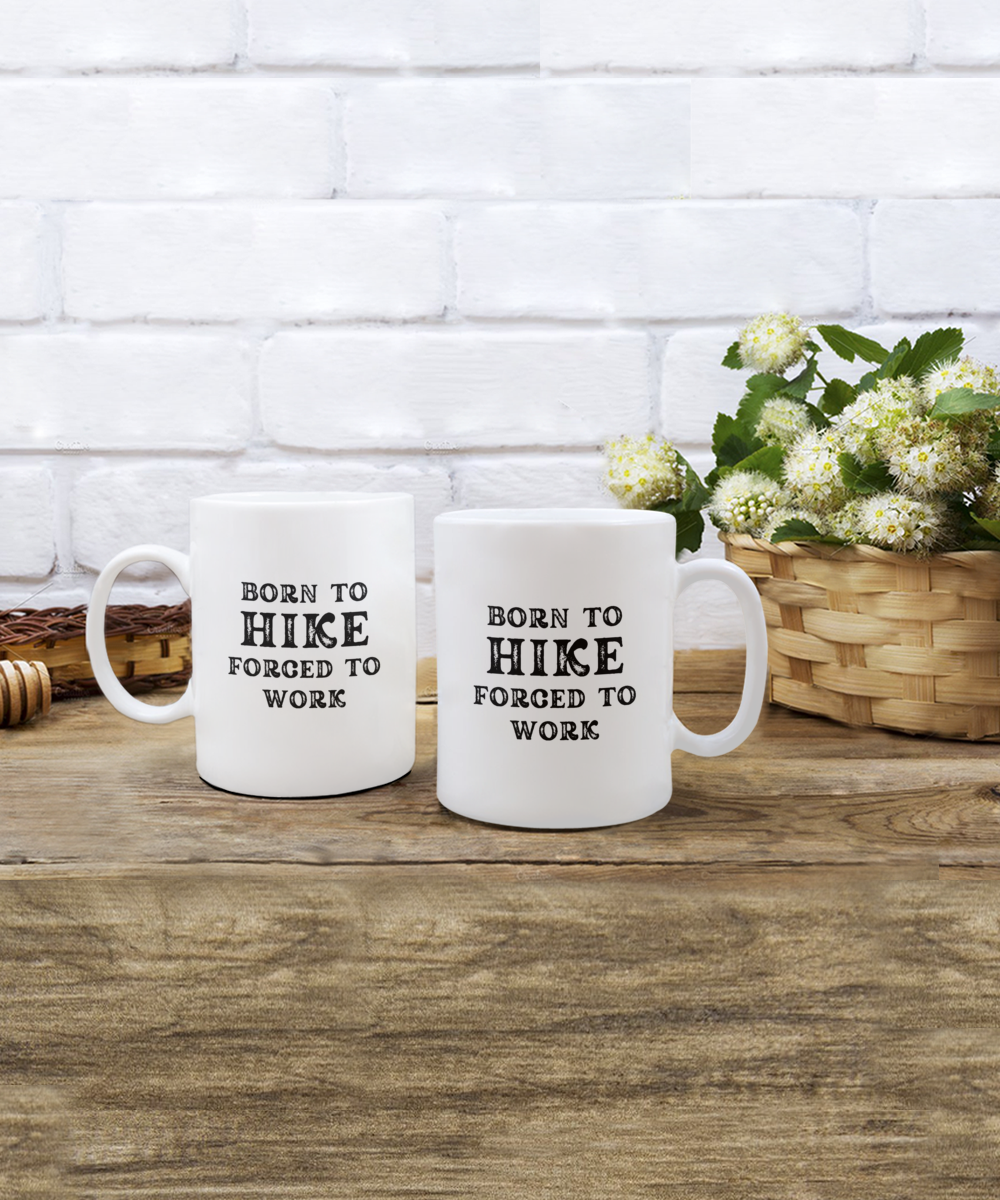 Two white "Born to Hike, Forced to Work" coffee mugs rest on a wooden table by white flowers. These charming mugs are printed and shipped in the USA, NOT store-available. Get your Born to Hike Forced to Work Coffee Mug, the perfect hiker gift, for a limited time!.