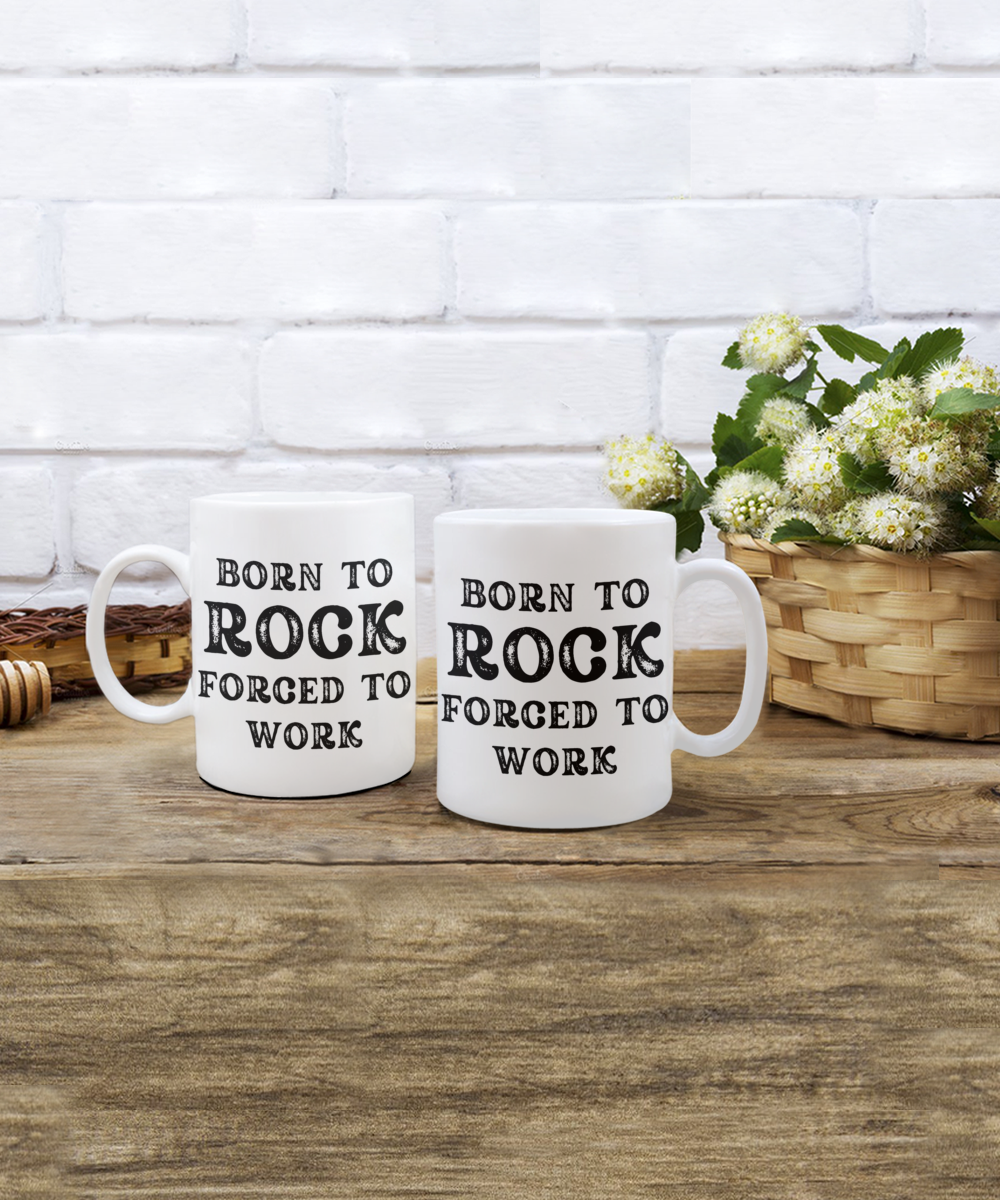 Two white "Born to Rock, Forced to Work" mugs sit on a wooden surface with a wicker basket of white flowers against a brick wall. These delightful mugs are printed and shipped from the USA and available for a limited time as the perfect gift for rock enthusiasts.